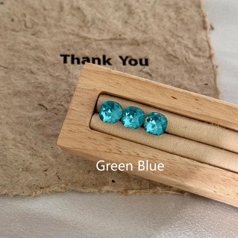 May include: Three square-cut, green-blue gemstones are displayed in a light wood box. The box sits on a textured, brown paper with the words "Thank You" printed in black. The text "Green Blue" is printed below the gemstones.