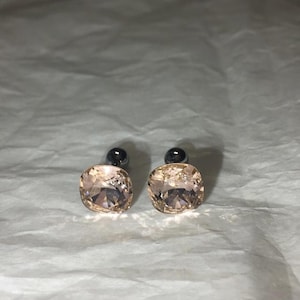 May include: A pair of stud earrings featuring large, square-cut, peach-colored crystals. The earrings have a silver-colored post and backing. The crystals are faceted, catching the light and creating a sparkling effect.
