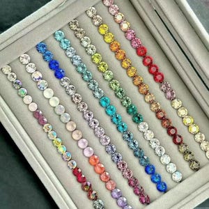 Puede incluir: Una exhibición de pulseras de pedrería coloridas dispuestas en una caja de joyería de terciopelo gris. Las pulseras muestran un espectro de colores, incluyendo transparente, azul, verde, amarillo, naranja, rojo y morado. Cada pulsera está compuesta por una sola fila de pedrería brillante.