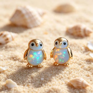 May include: A pair of stud earrings shaped like penguins, featuring gold-toned metal outlines and bodies made of iridescent white opal-like material with flecks of blue, green, and orange color throughout the surface of each penguin figure.