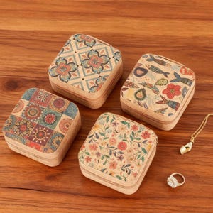 May include: Four square cork jewelry boxes with decorative patterns. The boxes feature floral, fish, and geometric designs in various colors. A gold necklace and ring are also visible on the wooden surface.