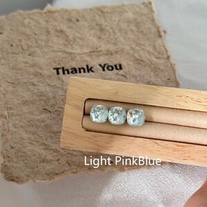 May include: Three light blue-green square gemstones are displayed in a wooden holder. The text "Light PinkBlue" is below the gemstones. A piece of textured paper with the words "Thank You" is in the background.