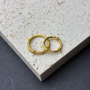 Puede incluir: Dos pendientes de aro dorados. Los pendientes son circulares con un diseño texturizado y retorcido. Los aros están entrelazados, uno ligeramente más grande que el otro. Los pendientes se muestran sobre una superficie clara y texturizada.