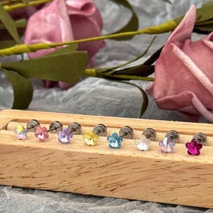 May include: A collection of flower-shaped stud earrings in various colors, including yellow, pink, purple, and blue. The earrings are displayed on a wooden block, with a blurred floral background.