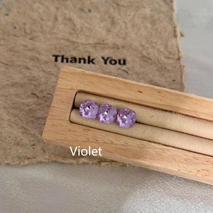 May include: Three violet-colored, square-cut gemstones displayed in a wooden jewelry holder. The holder is set against a backdrop of a brown paper with the words "Thank You" printed on it. The word "Violet" is printed below the gemstones.