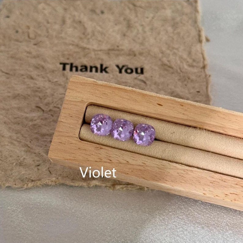 May include: Three violet-colored, square-cut gemstones displayed in a wooden jewelry holder. The holder is set against a backdrop of a brown paper with the words "Thank You" printed on it. The word "Violet" is printed below the gemstones.