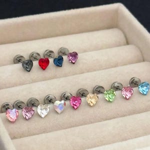 May include: A collection of heart-shaped stud earrings in various colors, including black, red, blue, pink, and iridescent. The earrings are displayed in a beige jewelry box, showcasing their polished metal posts and colorful crystal hearts.