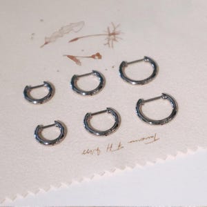 Puede incluir: Seis pendientes de aro plateados con cierre de bisagra se colocan sobre una superficie beige claro. Los pendientes circulares son pequeños y pulidos. El fondo presenta ilustraciones florales tenues y texto manuscrito.