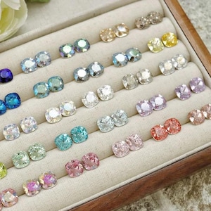 May include: A display of various colorful square-cut crystal stud earrings in a wooden jewelry box. The earrings are arranged in rows and include shades of blue, green, pink, and clear crystals. The box has a light-colored fabric lining.