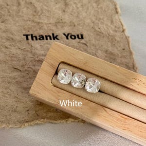 May include: Three white, square-cut gemstones are displayed in a light-colored wooden jewelry holder. The word "White" is printed in white below the gemstones. A brown paper with the words "Thank You" is in the background.