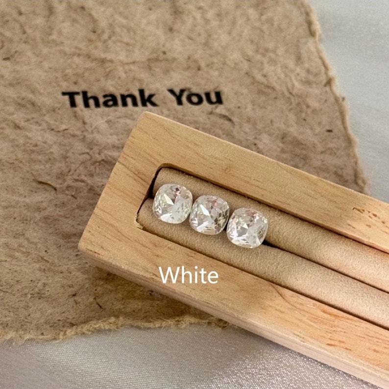 May include: Three white, square-cut gemstones are displayed in a light-colored wooden jewelry holder. The word "White" is printed in white below the gemstones. A brown paper with the words "Thank You" is in the background.