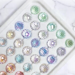 May include: A collection of iridescent, round, faceted glass gems in various colors, including green, blue, purple, and clear. The gems are arranged in a white tray, creating a sparkling and decorative display. These gems could be used for crafts or jewelry making.