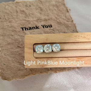 May include: Three light pink and blue square-shaped crystals are displayed in a wooden holder. The crystals are labeled "Light PinkBlue Moonlight". A piece of paper with the words "Thank You" is in the background.