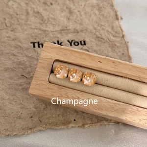 May include: Three champagne-colored square-cut gemstones are displayed in a wooden jewelry box. The box sits on a textured paper surface with the words "Thank You" visible. The word "Champagne" is printed below the gemstones.