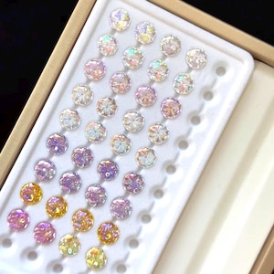 May include: A tray of iridescent, faceted gemstones in various colors, including clear, pink, purple, and yellow. The gemstones are arranged in a grid pattern within a white plastic insert, set inside a beige box.