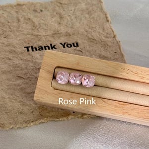 May include: Three rose pink cushion cut gemstones displayed in a wooden holder. The gemstones are a light pink color. The text "Rose Pink" is below the gemstones. A piece of brown paper with the text "Thank You" is in the background.