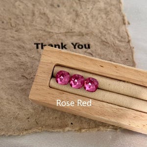 May include: Three rose red gemstones displayed in a wooden box. The gemstones are square-cut with a faceted surface, reflecting light. The box is set on a beige surface, with the words "Thank You" and "Rose Red" visible.