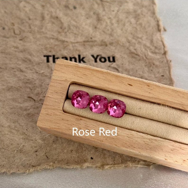 May include: Three rose red gemstones displayed in a wooden box. The gemstones are square-cut with a faceted surface, reflecting light. The box is set on a beige surface, with the words "Thank You" and "Rose Red" visible.