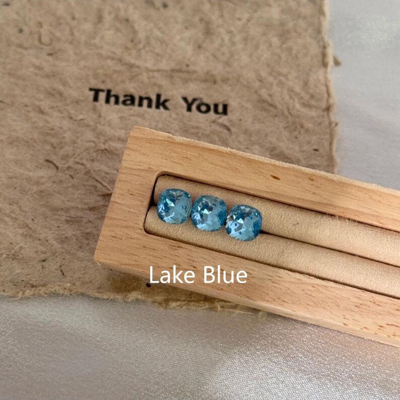 May include: Three faceted, lake blue gemstones are displayed in a wooden jewelry holder. The holder sits on a neutral-toned surface, with a piece of paper behind it that reads "Thank You". The text "Lake Blue" is printed below the gemstones.