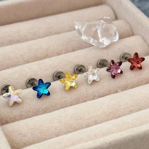 May include: A collection of six star-shaped stud earrings in various colors: iridescent, blue, yellow, clear, pink, and red. Each earring has a silver-colored post. The earrings are displayed in a beige jewelry box.