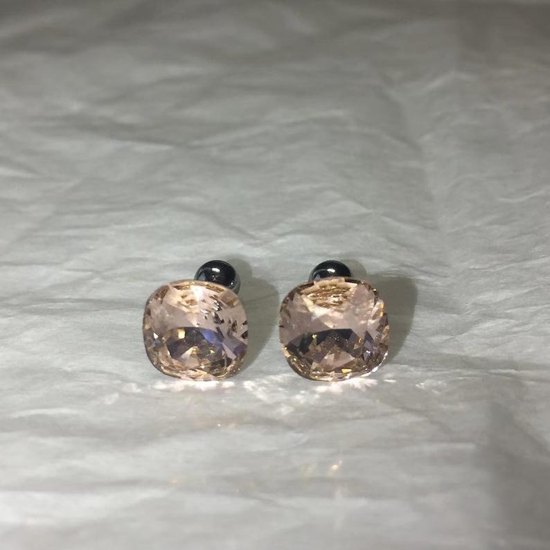 May include: A pair of stud earrings featuring square, peach-colored crystals. The crystals have a faceted design, reflecting light. Each earring has a small, dark gray post.