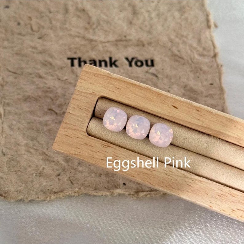 May include: Three square, light pink gemstones, labeled "Eggshell Pink", are displayed in a wooden tray. The tray sits on a textured, brown paper with the words "Thank You" printed on it.