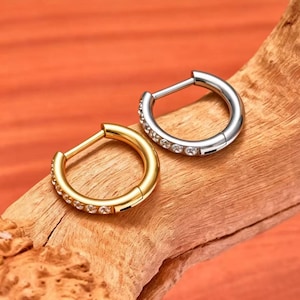 Puede incluir: Dos pendientes de aro, uno dorado y otro plateado, cada uno adornado con pequeñas gemas transparentes. Los pendientes se exhiben sobre un trozo de madera sobre un fondo naranja. El pendiente dorado tiene una fila de gemas.