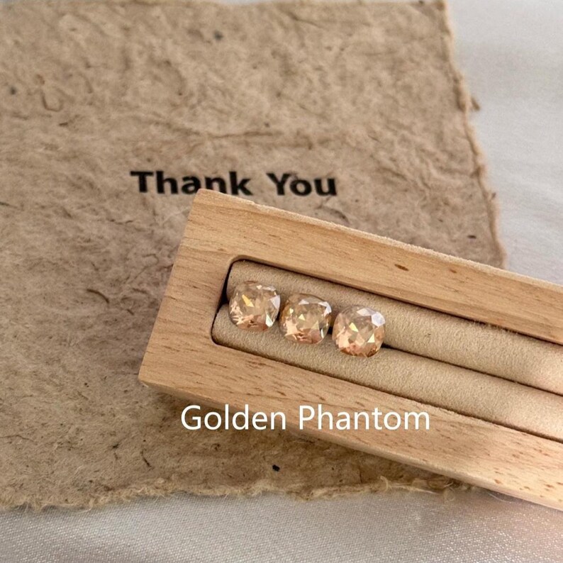 May include: Three golden-toned, square-cut gemstones are displayed in a light wooden box. The box sits on a textured, tan paper with the words "Thank You" printed on it. The words "Golden Phantom" are printed below the box.