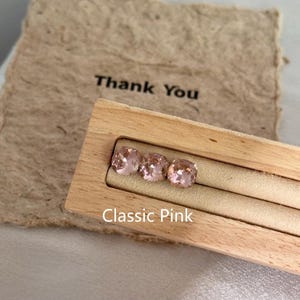 May include: Three square-cut, light pink gemstones displayed in a wooden holder. The gemstones are labeled "Classic Pink" and are set against a beige background. A beige paper with the words "Thank You" is in the background.