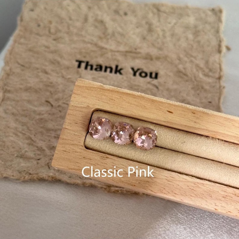 May include: Three square-cut, light pink gemstones displayed in a wooden holder. The gemstones are labeled "Classic Pink" and are set against a beige background. A beige paper with the words "Thank You" is in the background.