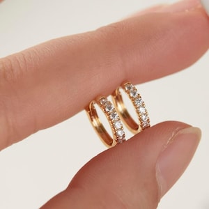 May include: A pair of gold hoop earrings, each adorned with a row of small, clear gemstones. The earrings are held between two fingers, showcasing their delicate design and sparkling details. The gold setting provides a warm contrast to the clear stones.