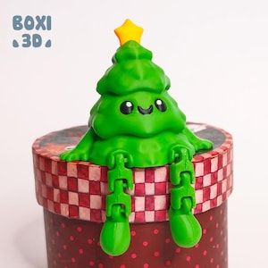May include: A green Christmas tree figurine with a yellow star on top, sitting on a red and white checked gift box. The tree has a smiling face and articulated legs.
