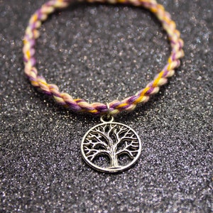 以下が含まれることがあります： 黄色、白、紫の糸で編まれた友情ブレスレットに、シルバーの生命の木のチャームが付いている。