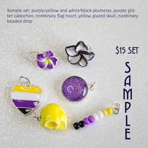 以下が含まれることがあります： 紫と黄色のプルメリアの花、白と黒のプルメリアの花、紫色のグリッターカボション、ノンバイナリーフラッグハート、黄色の釉薬で覆われたスカル、ノンバイナリービーズドロップの6つのチャームのセット。チャームはキラキラした表面に配置されています。$15 SET SAMPLE