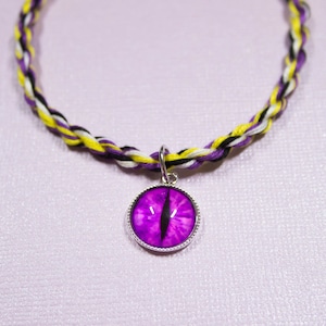 Könnte beinhalten: Eine geflochtene Halskette aus lila und schwarzem Kordel mit einem silbernen Anhänger, der ein lila Auge mit einer schwarzen Pupille zeigt.