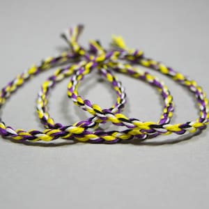 May include: Three braided bracelets in yellow, purple, black, and white colors. The bracelets are knotted together.