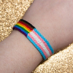 Puede incluir: Un brazalete de amistad tejido con los colores del arcoíris y la bandera del orgullo transgénero. El brazalete es azul, verde, amarillo, naranja, rojo, morado, negro, marrón, azul claro, rosa, blanco y azul claro.
