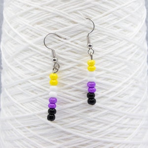 May include: A pair of dangle earrings with a silver hook and a beaded charm. The beads are yellow, white, purple, and black, representing the non-binary pride flag.