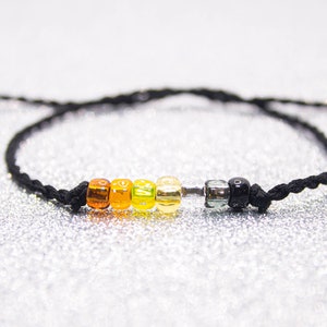 Könnte beinhalten: Ein Armband aus schwarzem Band mit einem Regenbogen aus bunten Perlen. Die Perlen sind orange, gelb, grün und klar. Am Ende des Armbands befindet sich eine schwarze Perle.