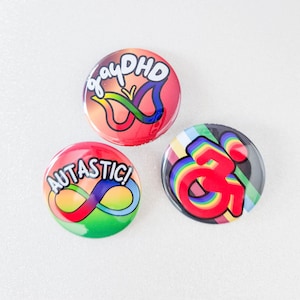 May include: Three round button pins with rainbow designs. The first pin has a red background with a butterfly and the text "gayDHD". The second pin has a green background with an infinity symbol and the text "AUTASTIC!". The third pin has a black background with a rainbow-colored wheelchair symbol.