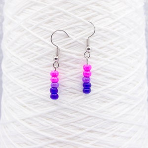 May include: A pair of dangle earrings with a gradient of pink, purple, and blue beads. The earrings are made with silver hooks.