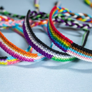 Pode incluir: Cinco pulseiras de amizade coloridas com diferentes padrões de bandeiras do orgulho, incluindo as bandeiras do arco-íris, genderqueer e assexual.
