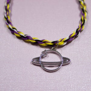Op de afbeelding: Een gevlochten ketting met gele, witte, zwarte en paarse draden. Een zilveren hanger hangt aan de ketting, die een planeet met een ring en een kleine ster voorstelt.