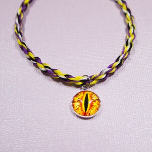 May include: A yellow, white, and purple braided cord necklace with a silver charm featuring a yellow eye with a black pupil.