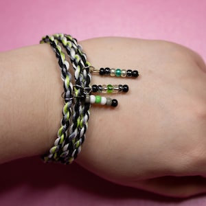 May include: A black, white, and green braided bracelet with three beaded charms. The charms are black, white, and green beads.
