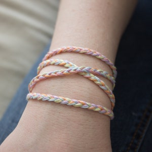 Puede incluir: Un brazalete trenzado multicolor hecho de hilo rosa pastel, amarillo y azul. El brazalete se lleva en la muñeca.