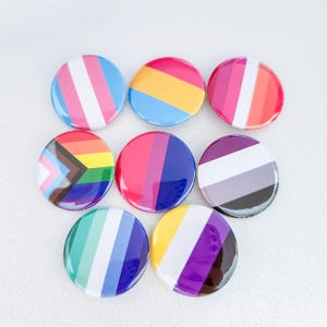 May include: Nine round buttons with different pride flags printed on them. The flags include the transgender flag, the pansexual flag, the lesbian flag, the bisexual flag, the asexual flag, the genderfluid flag, the non-binary flag, the intersex flag, and the gay pride flag.