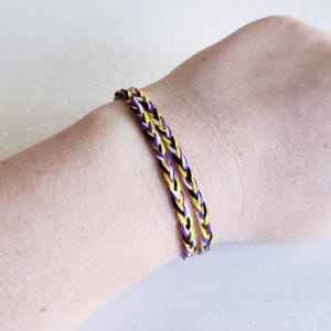 Pode incluir: Duas pulseiras de amizade trançadas nas cores amarelo, branco e roxo. As pulseiras são usadas no pulso.
