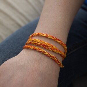 May include: A close-up of a person's wrist wearing three braided bracelets. The bracelets are made of yellow and orange thread.