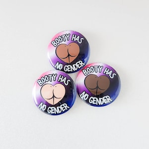 Puede incluir: Tres botones redondos con un degradado de colores morado, rosa y azul. Cada botón presenta un contorno en forma de corazón con una silueta desnuda, marrón y negra en el interior. El texto en cada botón dice "Booty Has No Gender".